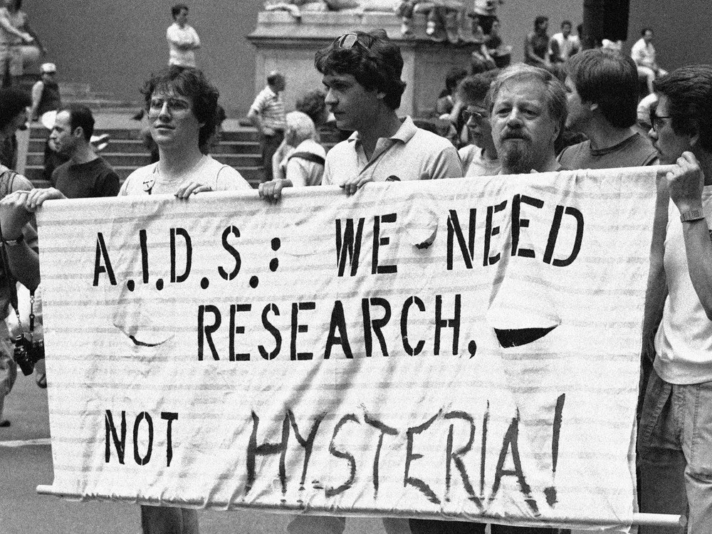 An image of three men holding up a sign that reads: A.I.D.S: WE NEED RESEARCH, NOT HYSTERIA.